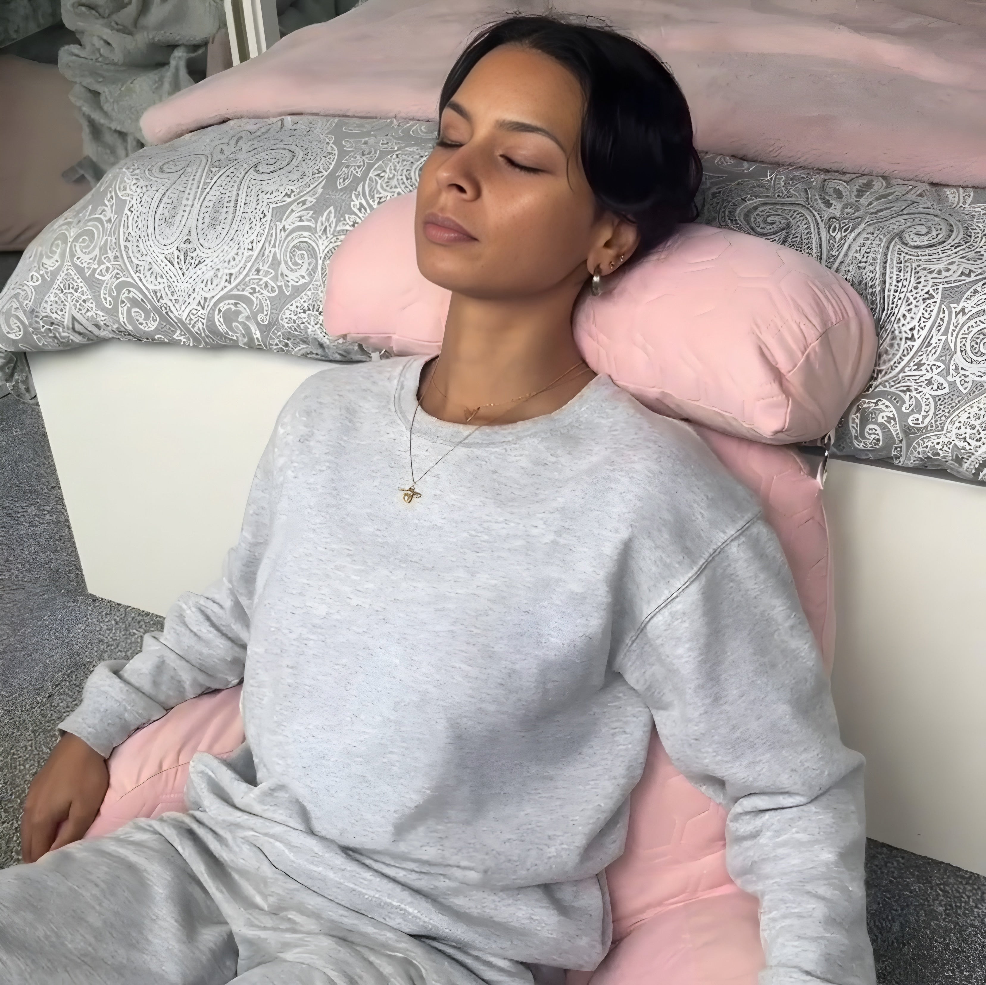 Woman relaxing on a soft pink ergonomic cushion with a detachable headrest, seated in a cosy space with grey bedding in the background. The cushion provides ultimate back and neck support, perfect for comfort and relaxation